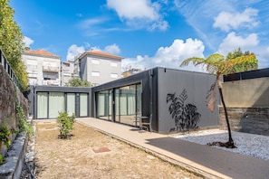 Un bâtiment moderne doté de grandes fenêtres en verre, d’une façade métallique et d’un petit espace jardin agrémenté d’un arbre et d’un chemin de gravier.
