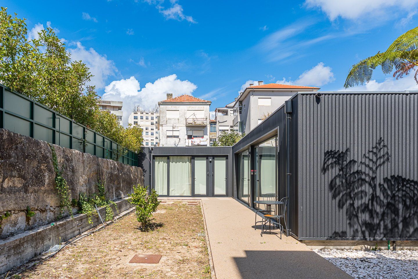 Un patio con un edificio moderno, un muro de piedra y un pequeño árbol.