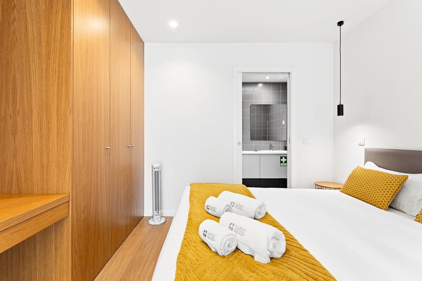 Une chambre d’hôtel moderne avec une armoire en bois, un lit recouvert de draps blancs et d’un plaid jaune, une table de chevet et une salle de bain visible à travers une porte ouverte.