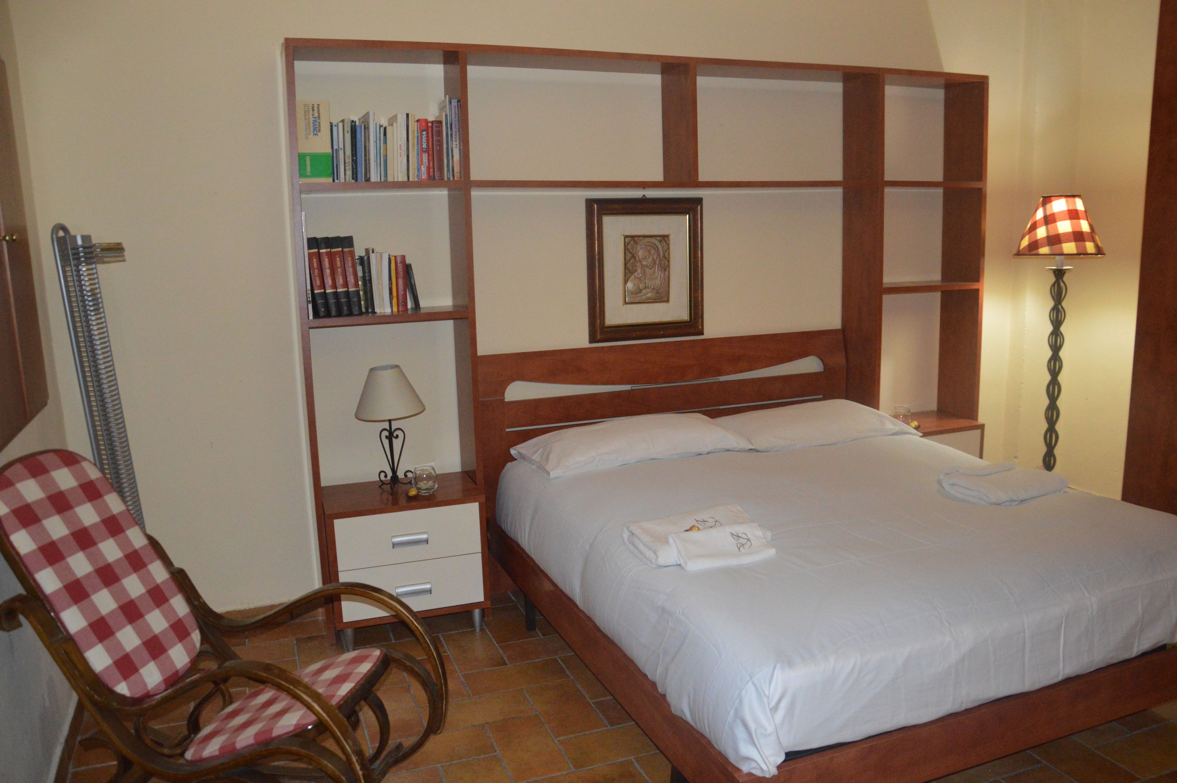 A hotel room with a bed, a chair, a bedside table with a lamp, a bookshelf, and a framed picture on the wall.