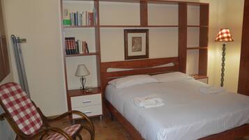 A hotel room with a bed, a chair, a bedside table with a lamp, a bookshelf, and a framed picture on the wall.