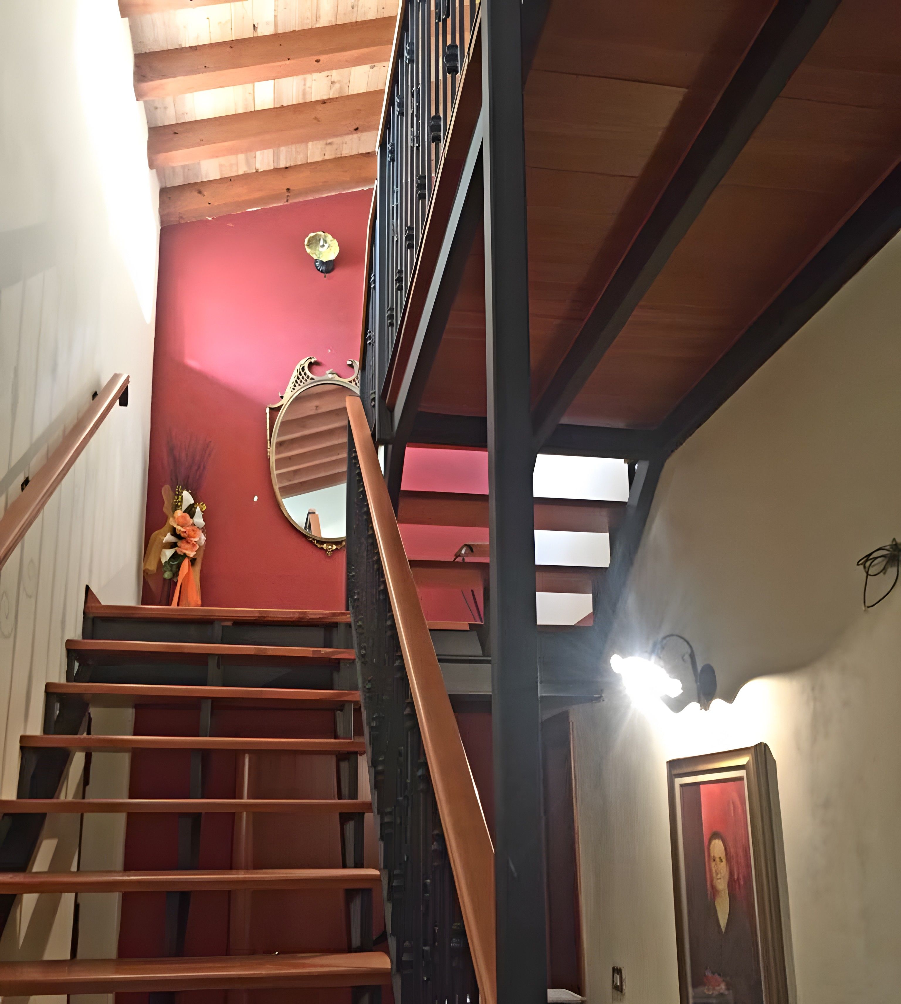 A staircase with wooden steps and a black handrail, leading to a red wall with a decorative mirror and a framed picture.