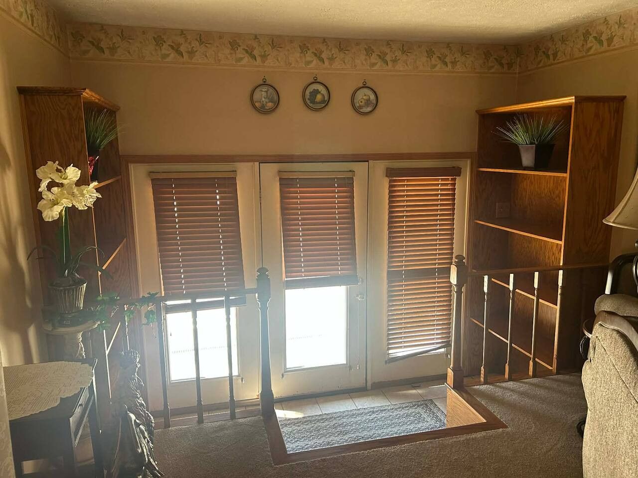 Una habitación con escalera de madera, una puerta con paneles de vidrio y una pequeña mesa con un florero.
