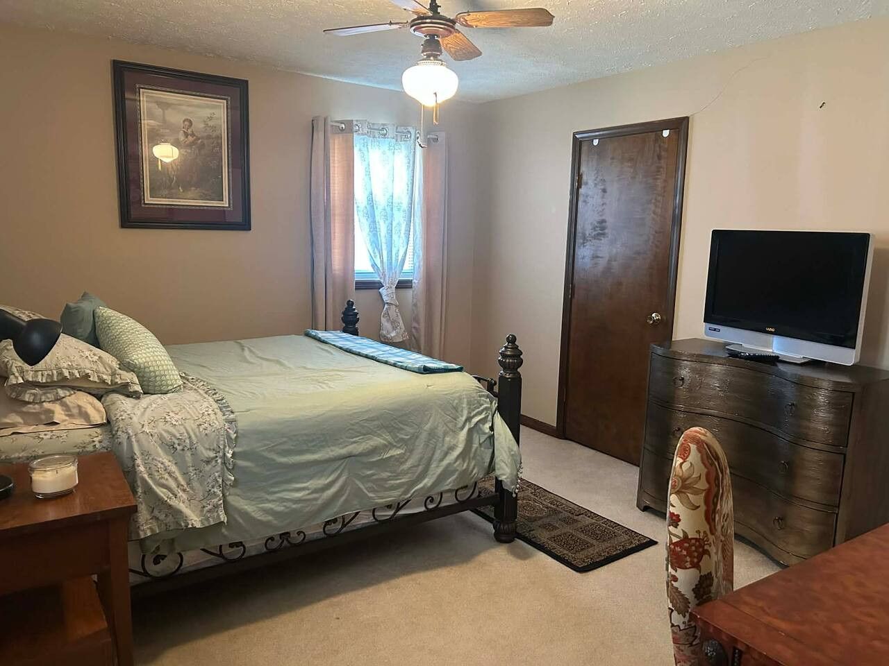 A bedroom with a bed, a television on a stand, a ceiling fan, a door, and a window with curtains.
