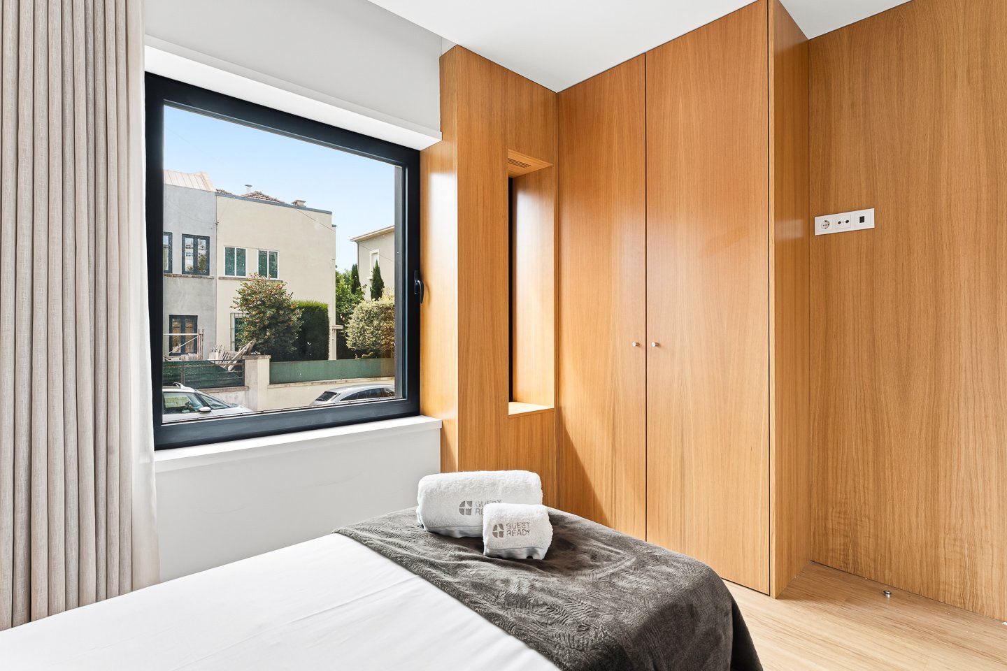 A modern bedroom with a large window, wooden wardrobe, and a bed with a grey blanket and two towels.