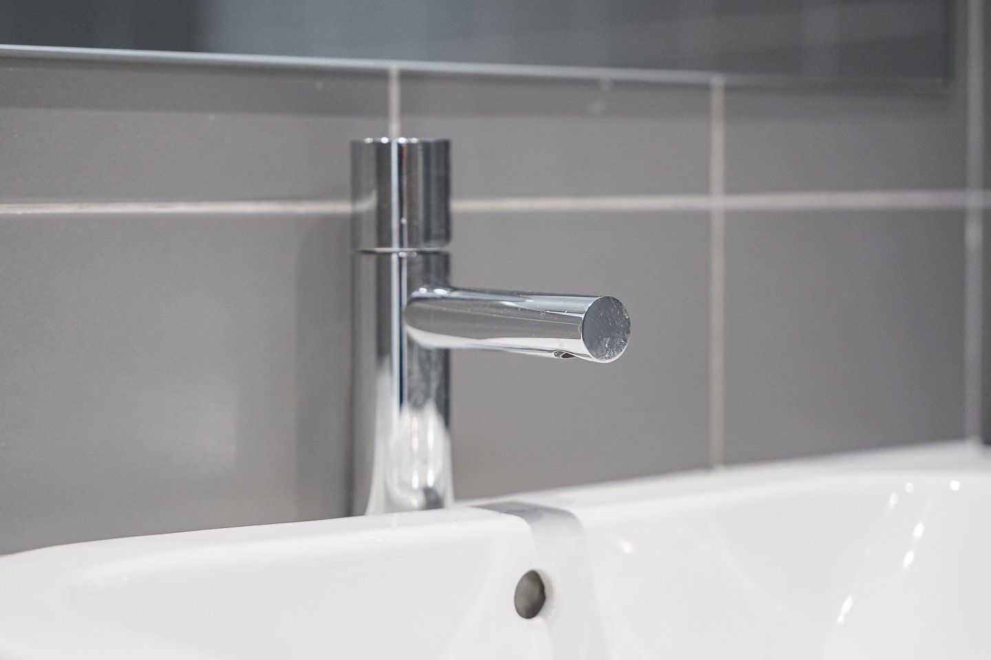 Ein chromfarbener Wasserhahn an einer weißen Badewanne in einem Badezimmer mit grauen Fliesenwänden.