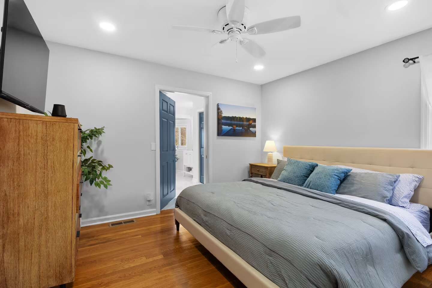 Un dormitorio con cama, ventilador de techo, mesita de noche, lámpara y vista a un baño a través de una puerta abierta.
