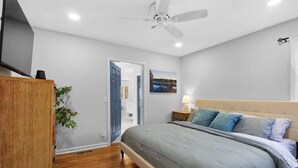 Ein Schlafzimmer mit einem Bett, einem Deckenventilator, einem Nachttisch, einer Lampe und Blick durch eine offene Tür auf ein Badezimmer.