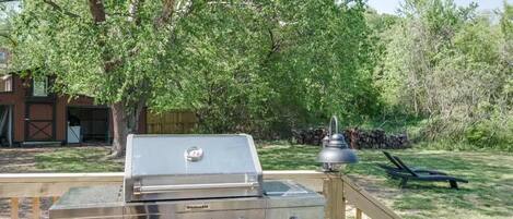 A stainless steel outdoor grill with four burners, a chimney, and a control panel, set against a wooden fence and surrounded by trees.