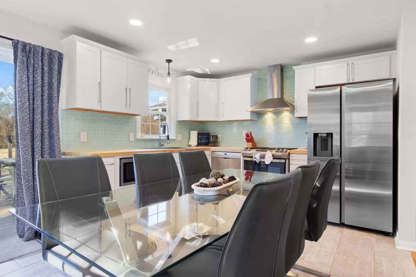 A modern kitchen with white cabinets, a stainless steel refrigerator, and a glass dining table with chairs.