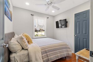 A bedroom with a bed, a ceiling fan, a TV mounted on the wall, a window with curtains, and a door. - *NEW* Shoreline Escape: Lakefront Retreat w/ Hot Tub (Winchester)