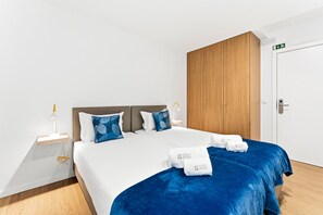 A hotel room with a bed, two pillows, a nightstand, a lamp, a wooden wardrobe, and a door with an emergency exit sign. - GuestReady - PoetikBlue - Room 1 (Porto)