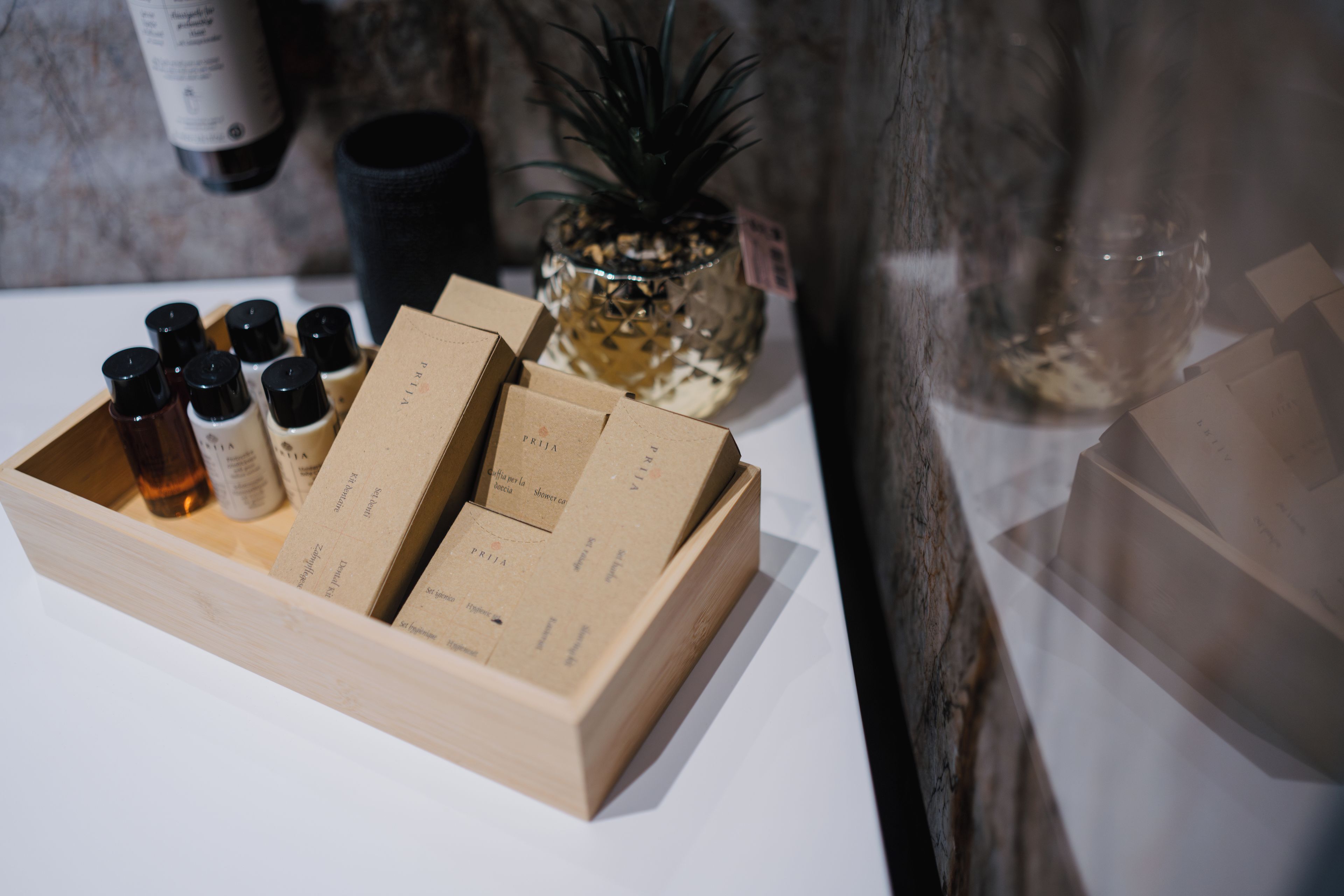 Une boîte en bois contenant plusieurs bouteilles et des boîtes étiquetées, probablement des articles de toilette ou des produits d’hygiène personnelle, est posée sur une table. On aperçoit une plante et une bouteille à l’arrière-plan.