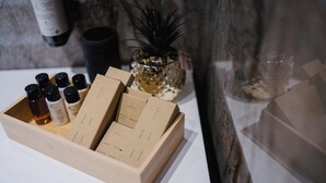 A wooden box containing several bottles and labeled boxes, likely toiletries or personal care items, on a table with a plant and a bottle in the background. - Harry's House Trani (Trani)