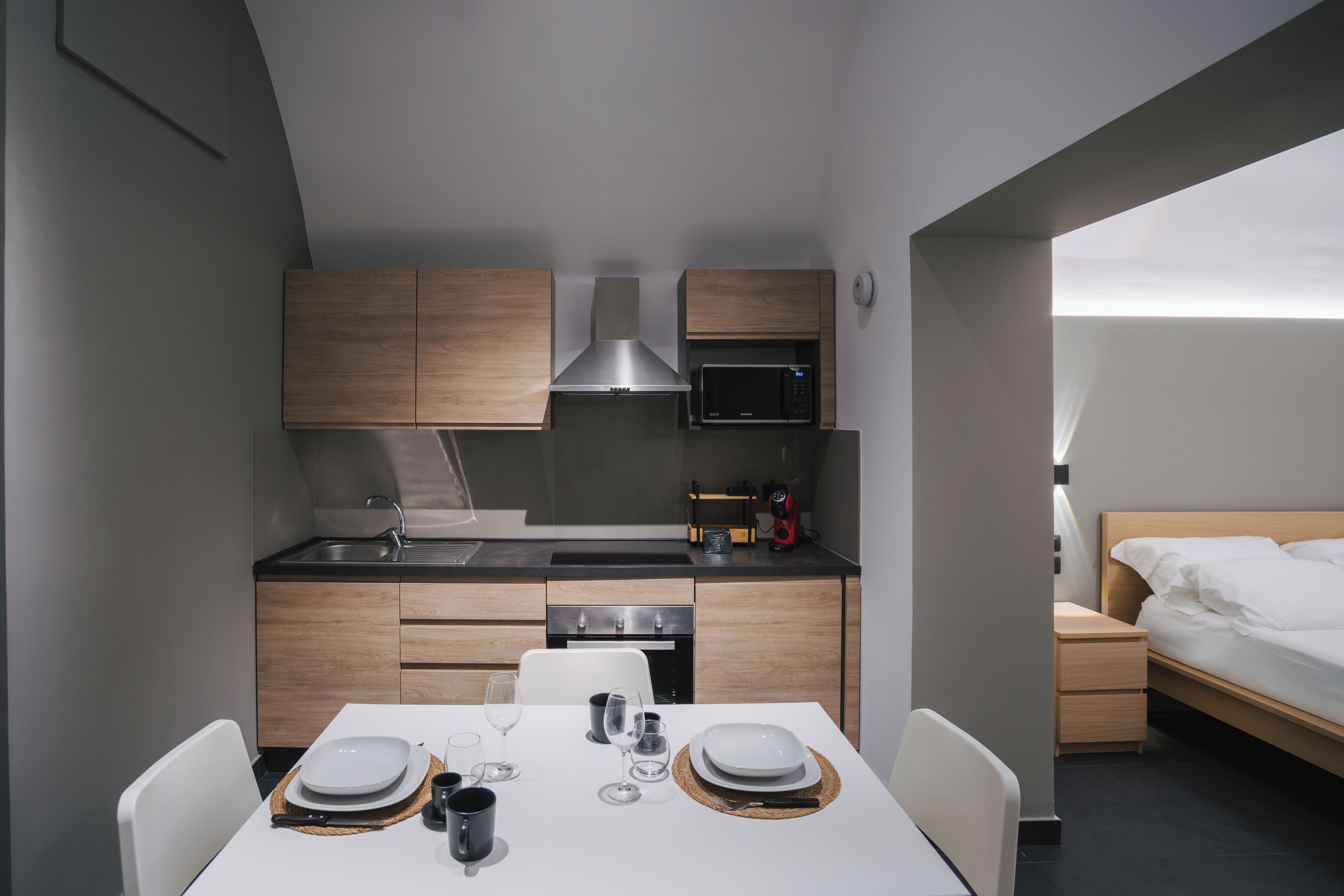 Une cuisine compacte équipée d’appareils modernes, un coin repas avec une table dressée pour deux personnes, et un lit dans la pièce adjacente.