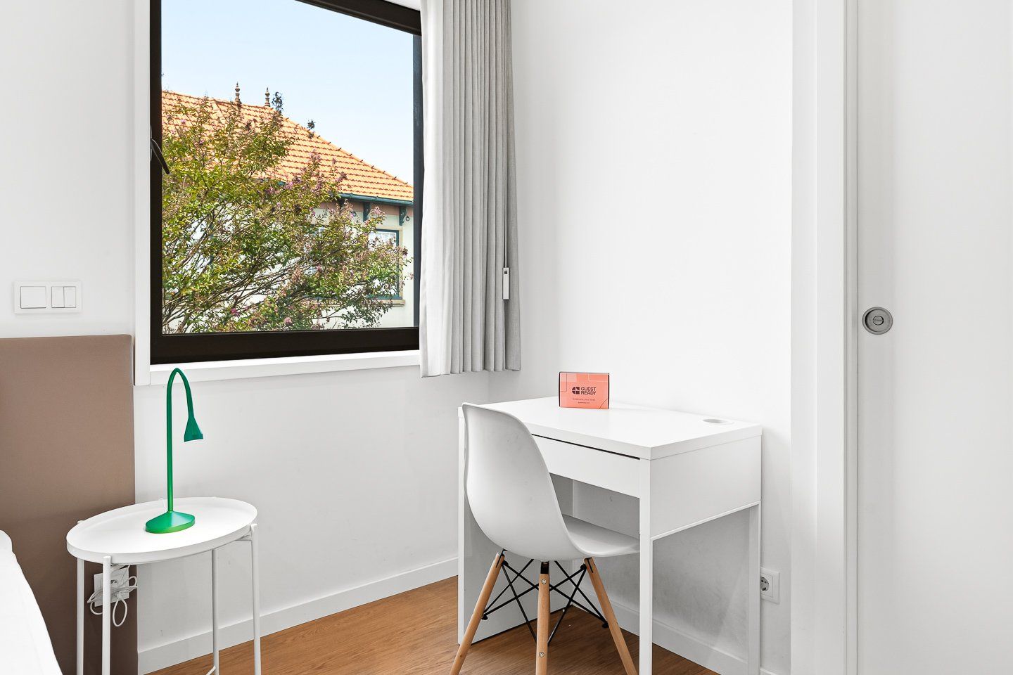 Ein moderner, minimalistischer Raum mit weißem Schreibtisch, einer grünen Schreibtischlampe und Blick auf ein Gebäude durch ein Fenster.