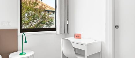 Ein moderner, minimalistischer Raum mit weißem Schreibtisch, einer grünen Schreibtischlampe und Blick auf ein Gebäude durch ein Fenster.
