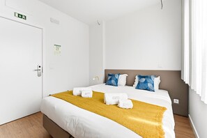 Quarto de hotel com cama, cabeceira, janela com cortinas, porta e luminária fixada na parede.