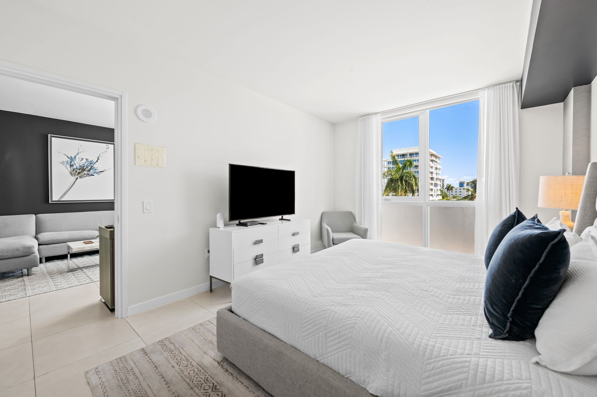 Quarto de hotel moderno com uma cama grande, televisão de tela plana, área de estar com sofá e poltrona, e vista para a cidade pelas janelas.