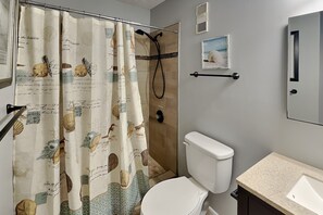 Casa de banho com cortina de chuveiro com motivos náuticos, sanita branca, lavatório com bancada e um quadro na parede.