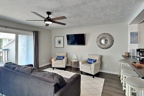 A modern living room with a grey sofa, two armchairs, a ceiling fan, a flat-screen TV, decorative wall art, and a kitchen area with bar stools. - Sandal Suite (Carolina Beach)