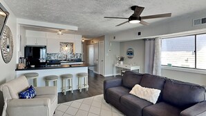 Una sala moderna con un sofá de cuero oscuro, un sillón de color claro y un tapete estampado. Se ve una cocina con armarios blancos y encimera negra al fondo.