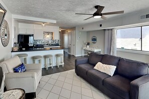 Una sala moderna con un sofá de cuero oscuro, un sillón de color claro y un tapete estampado. Se ve una cocina con armarios blancos y encimera negra al fondo.