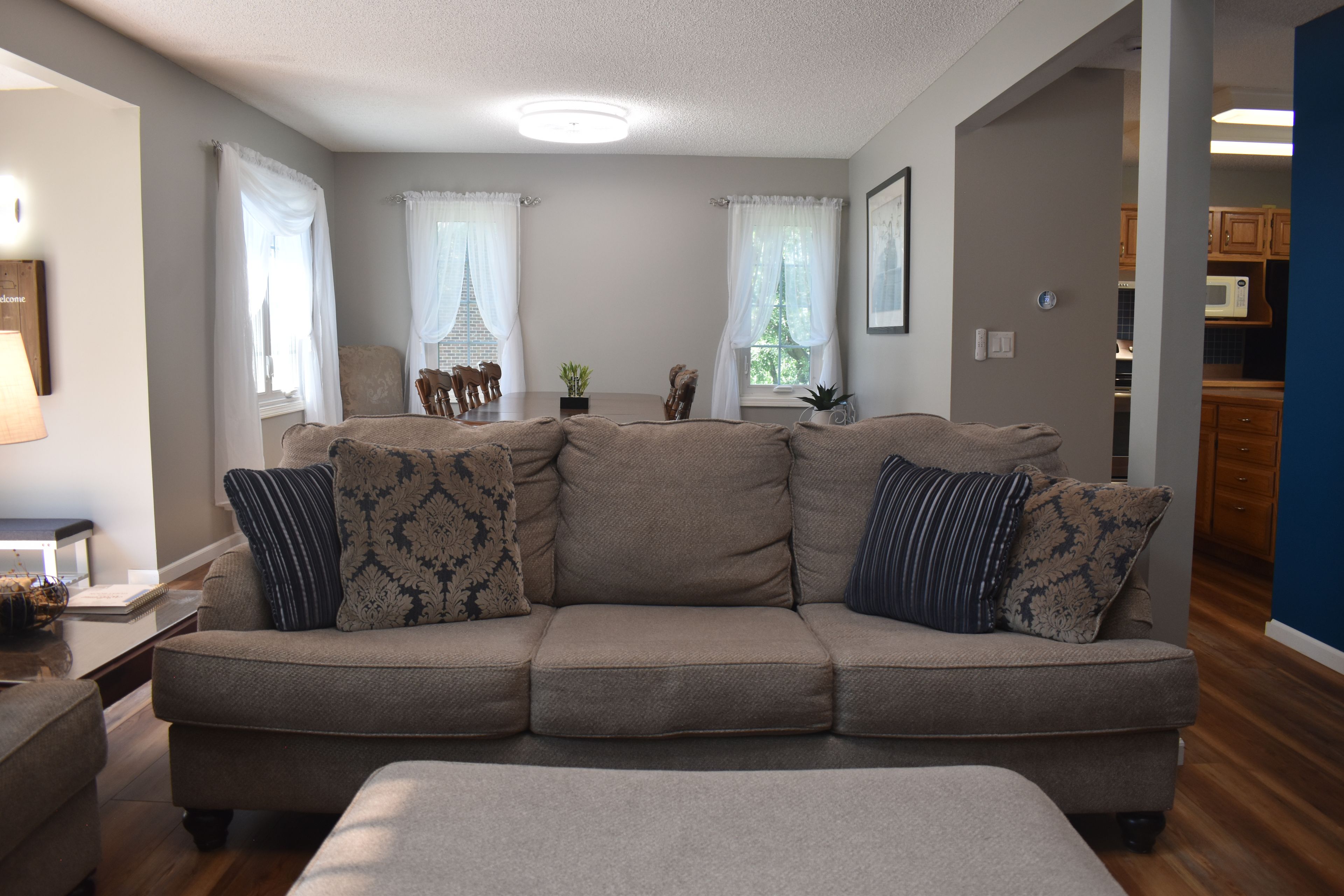 A living room with a beige sectional sofa, a small table, and a lamp. There are two windows with sheer curtains, a plant on the windowsill, and a framed picture on the wall.