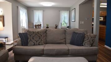 A living room with a beige sectional sofa, a small table, and a lamp. There are two windows with sheer curtains, a plant on the windowsill, and a framed picture on the wall.