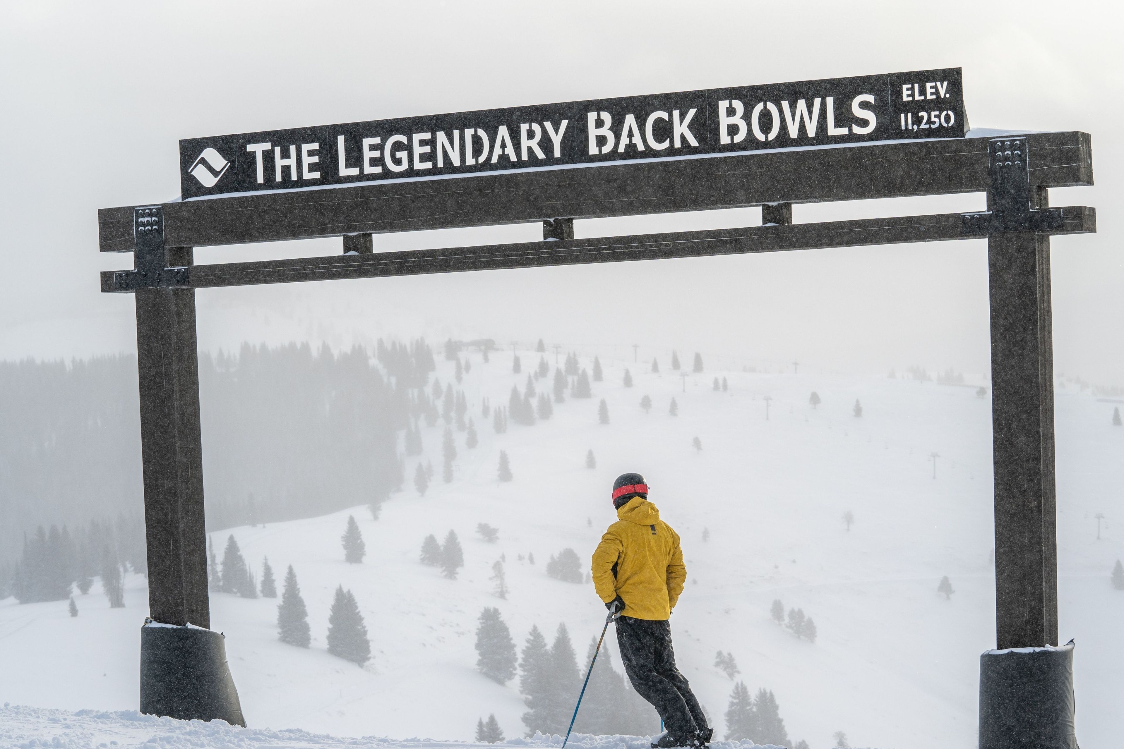 A ski slope with a sign reading 'The Legendary Back Bowls' and a skier in a yellow jacket.