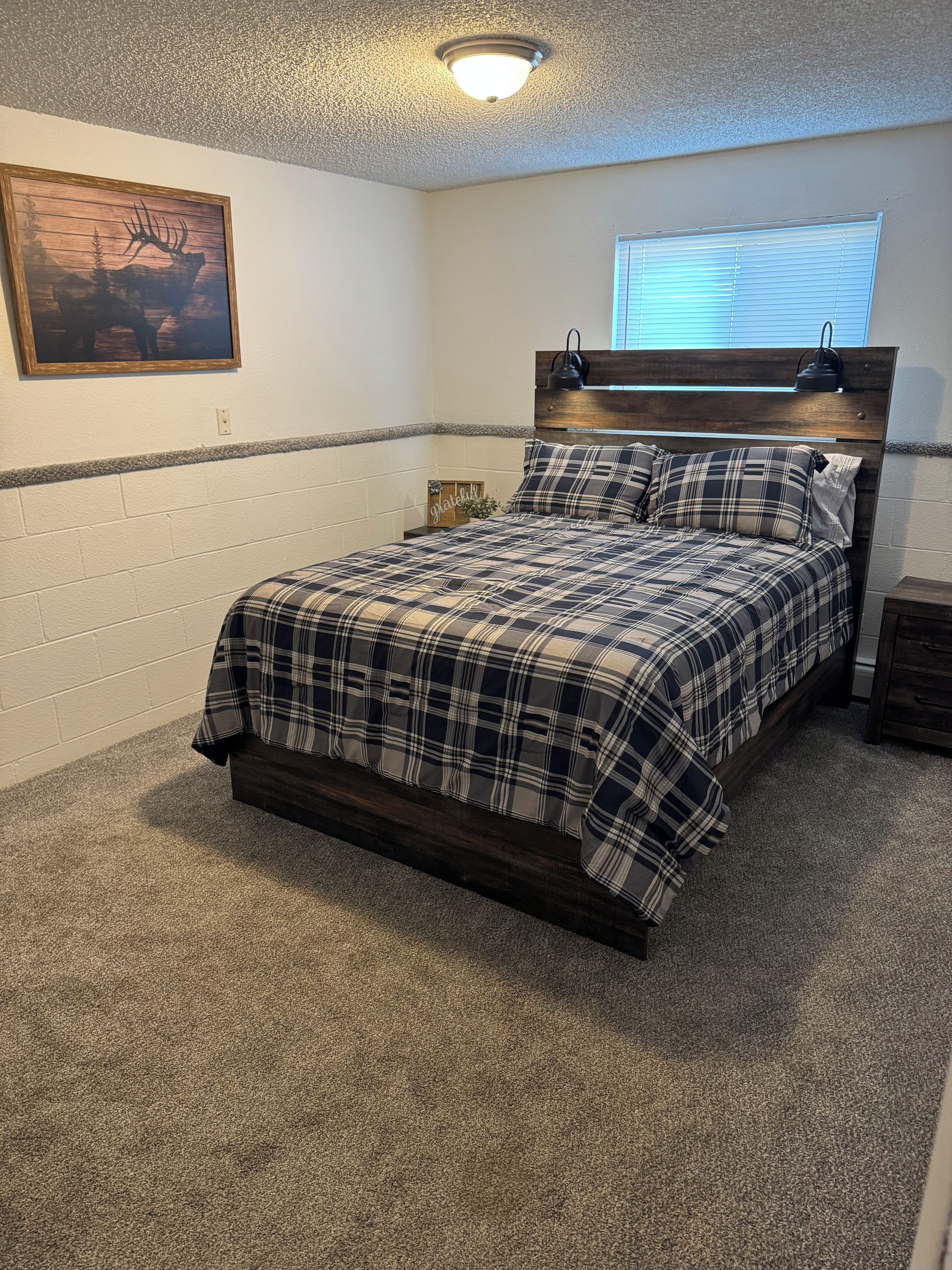 A bedroom with a bed, a headboard, a nightstand, a wall-mounted lamp, a window, and a framed picture.