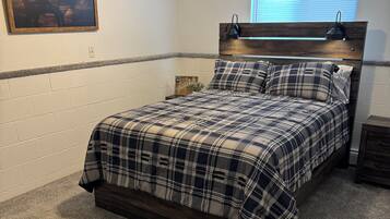 A bedroom with a bed, a headboard, a nightstand, a wall-mounted lamp, a window, and a framed picture.