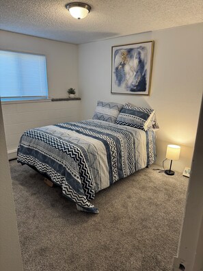 Ett sovrum med en säng, en vägglampa, ett fönster med persienner och en tavla på väggen.