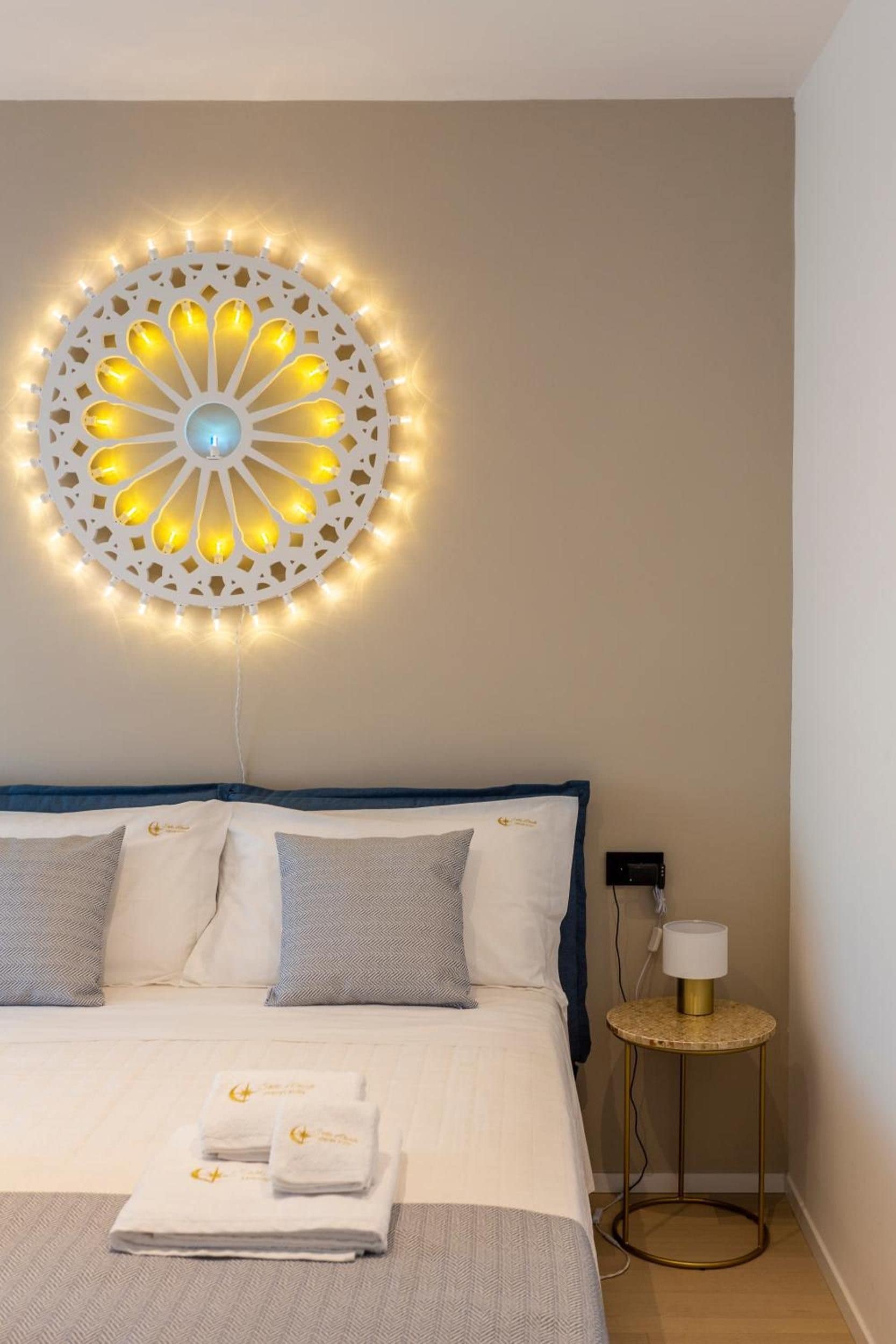 A hotel room with a bed, pillows, a wall-mounted light fixture, a bedside table with a lamp, and a folded towel on the bed.