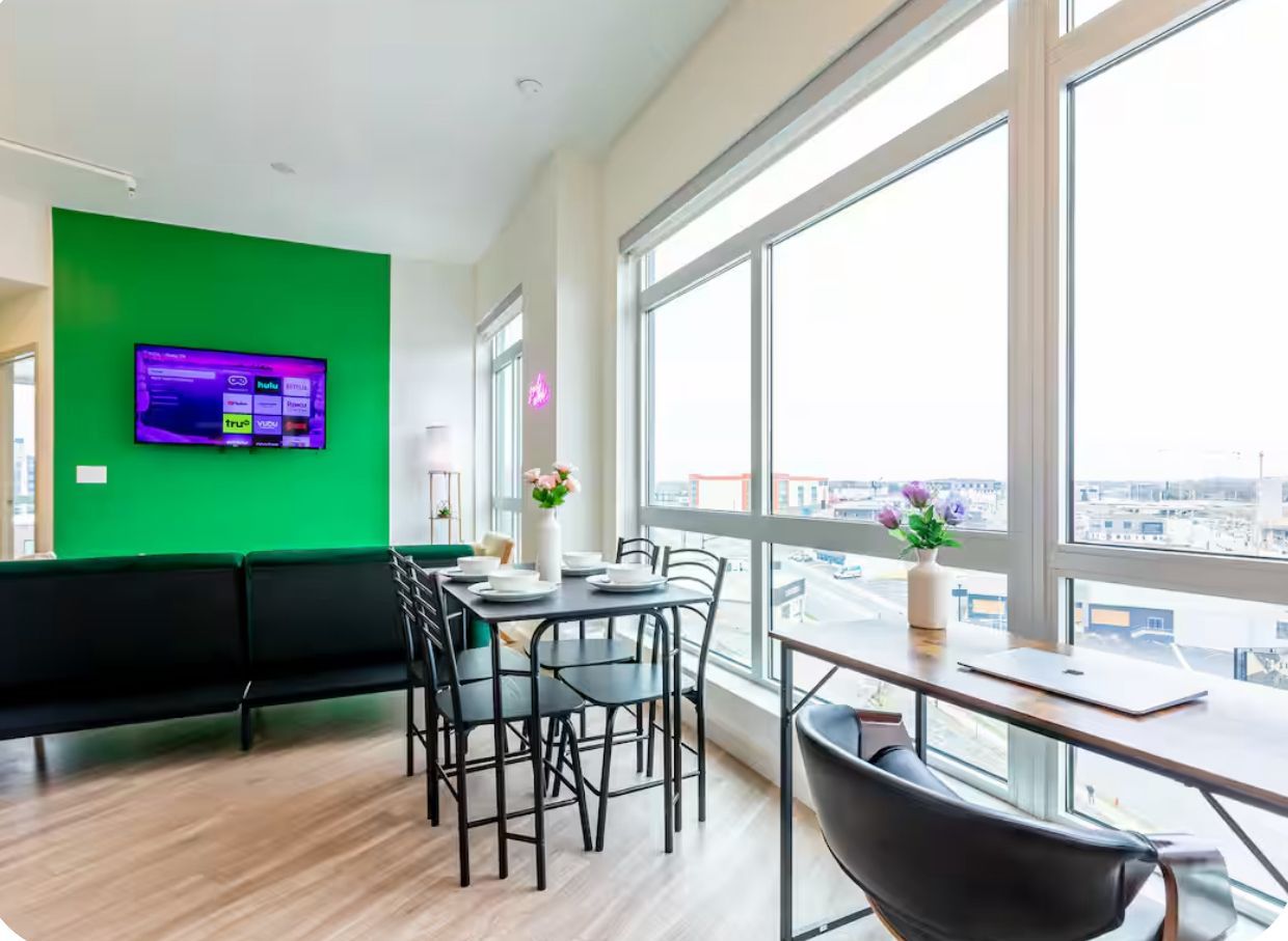 A modern interior with a green wall, a flat-screen TV, a dining area with a table and chairs, and large windows offering a city view.