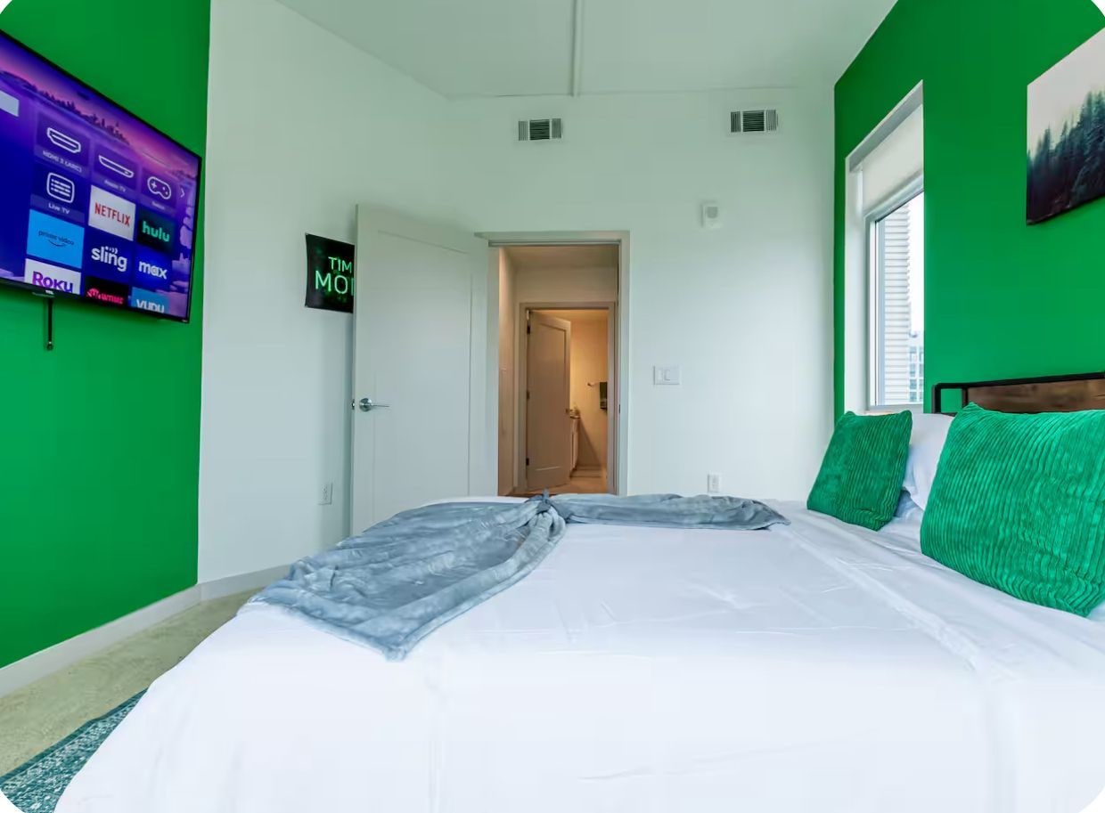 A room with a bed, green walls, a television displaying streaming services, and a door leading to another room.