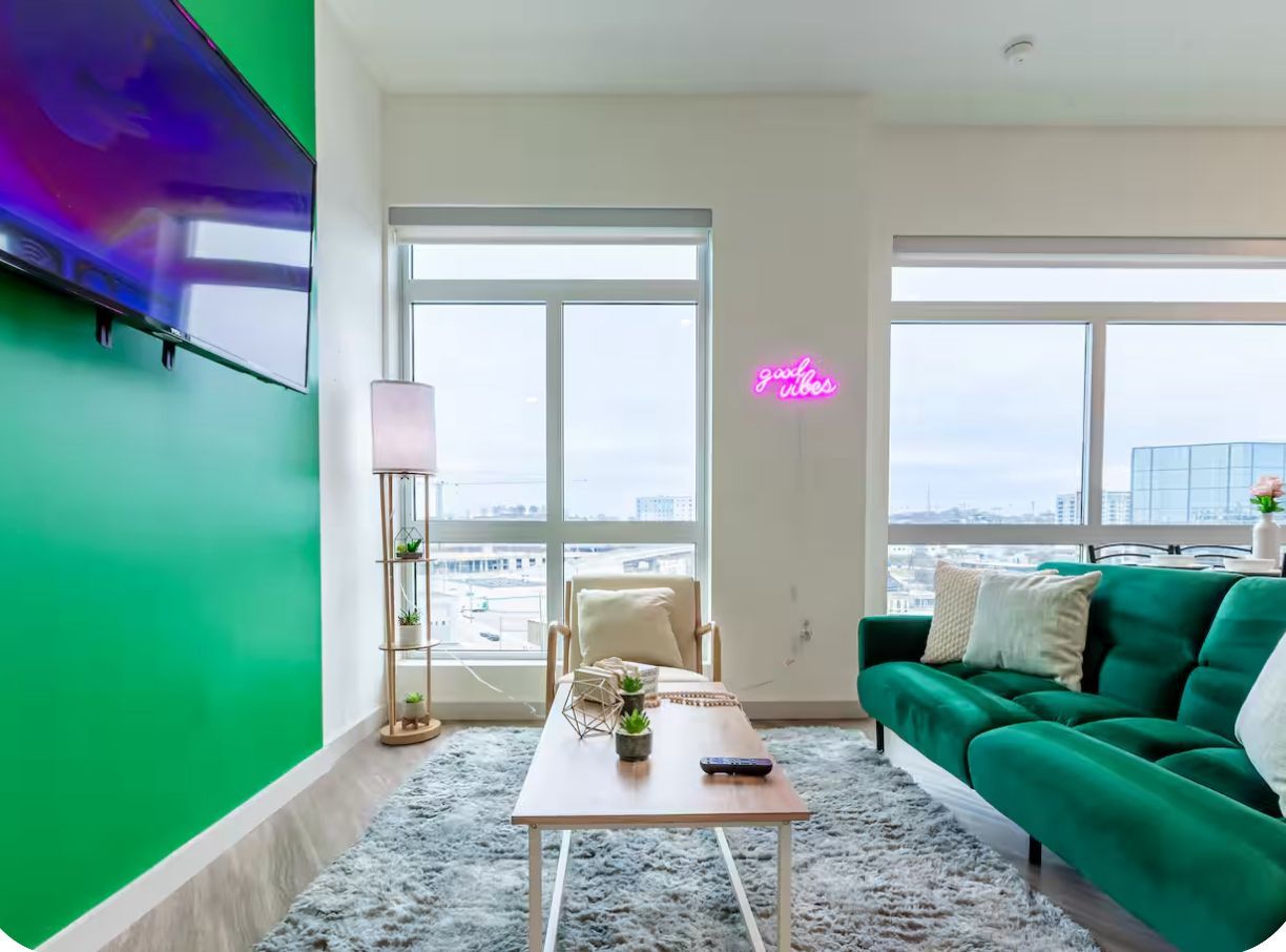 A modern living room with a green couch, a wooden coffee table, and a chair. There are two large windows providing a view of the cityscape.