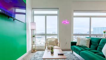 A modern living room with a green couch, a wooden coffee table, and a chair. There are two large windows providing a view of the cityscape.