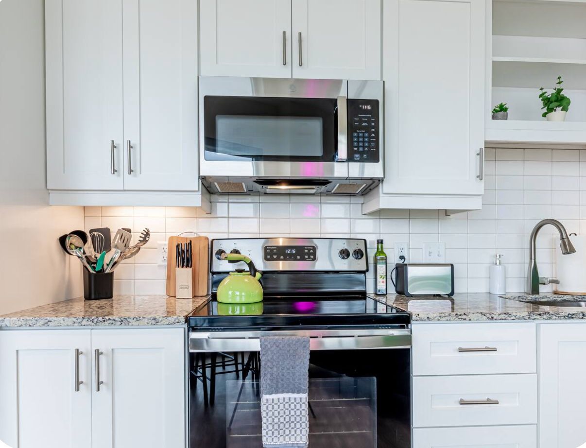 A modern kitchen with white cabinets, a stainless steel microwave, a green kettle, and a built-in oven.