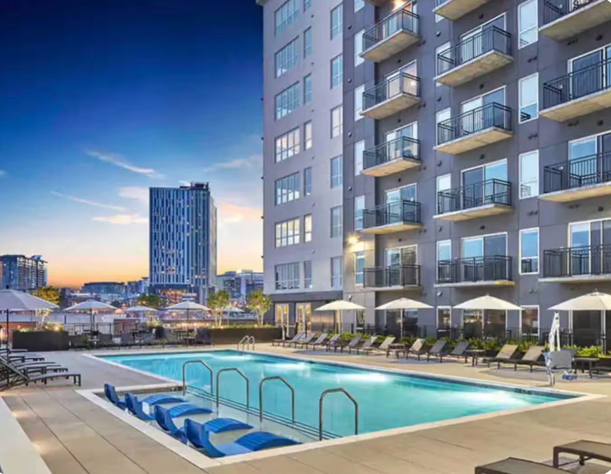 A rooftop pool area with lounge chairs and umbrellas, surrounded by a high-rise building with balconies.