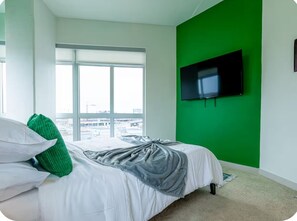 Un dormitorio con una cama, una pared verde, un televisor montado en la pared y una ventana con vistas a edificios.