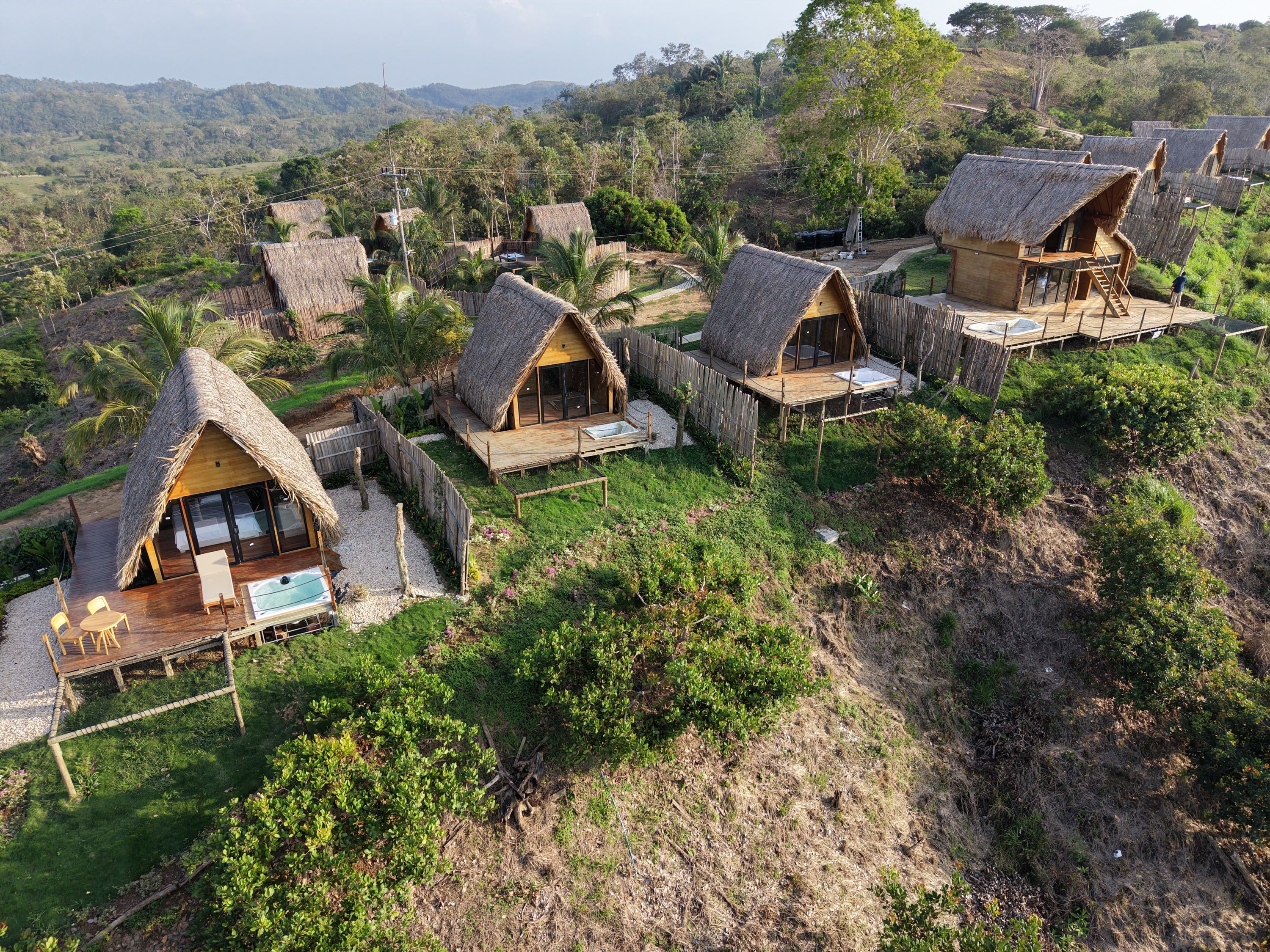 A cluster of thatched-roof cottages with wooden decks, surrounded by lush greenery and hills in the background.