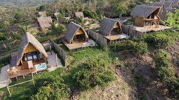 A cluster of thatched-roof cottages with wooden decks, surrounded by lush greenery and hills in the background.