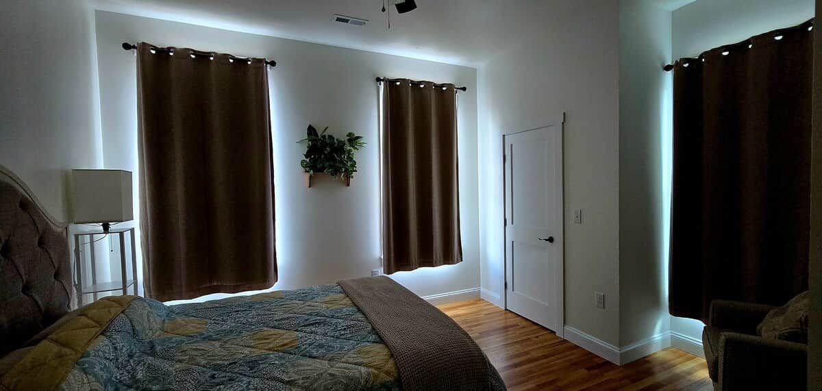 Un dormitorio con cama, dos ventanas con cortinas, una mesita de noche, una silla y una puerta.