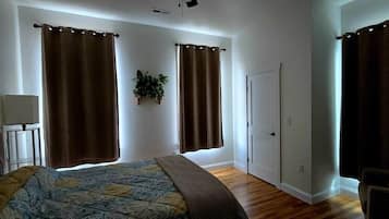 Un dormitorio con cama, dos ventanas con cortinas, una mesita de noche, una silla y una puerta.