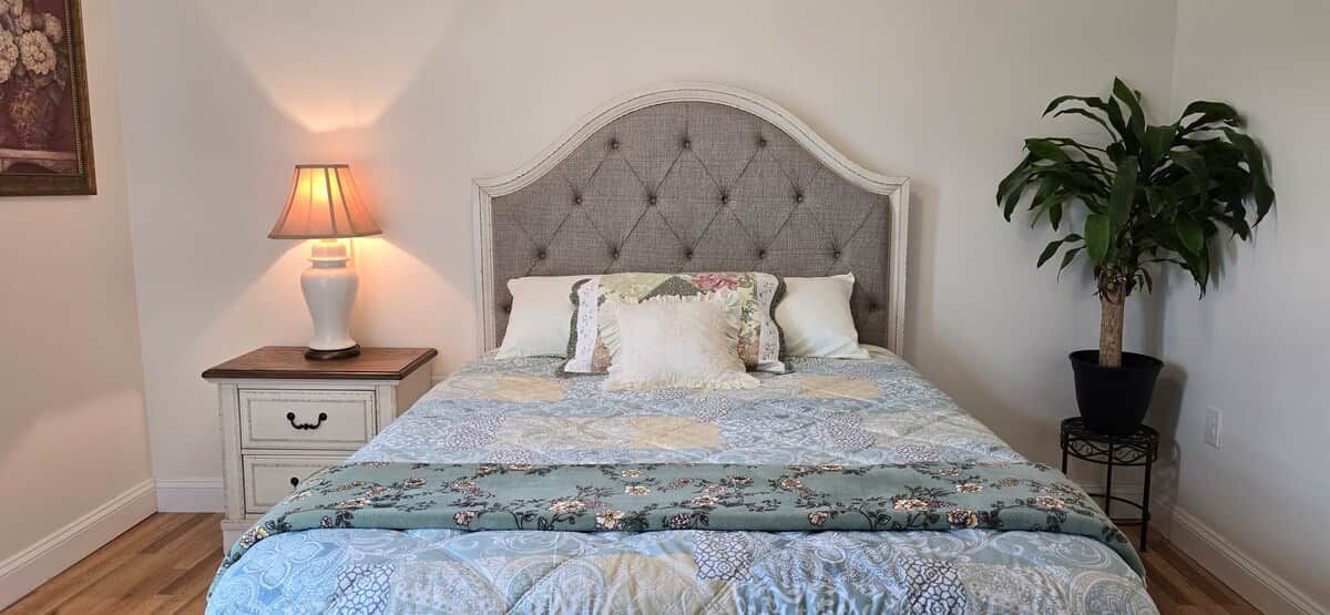 A bedroom with a bed, a nightstand with a lamp, a potted plant, and a wall-mounted shelf.