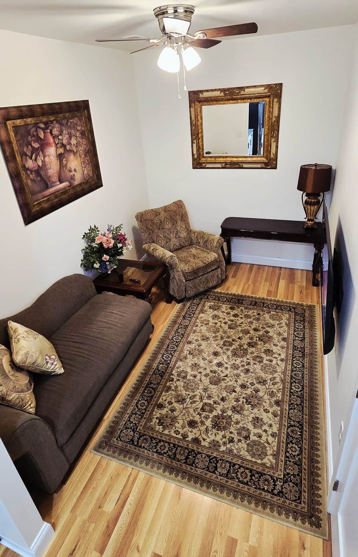 A living room with a brown sofa, a patterned armchair, a wooden coffee table, a large rug, a ceiling fan, a framed picture, and a lamp.