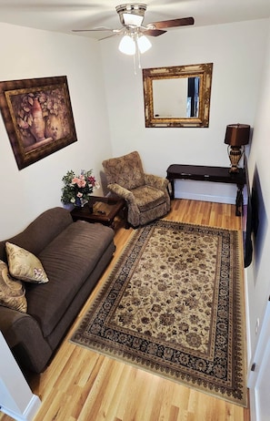 A living room with a brown sofa, a patterned armchair, a wooden coffee table, a large rug, a ceiling fan, a framed picture, and a lamp.