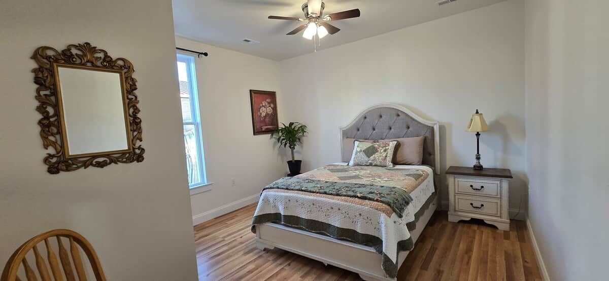A bedroom with a bed, a nightstand, a chair, a ceiling fan, a window, and a wall-mounted mirror.