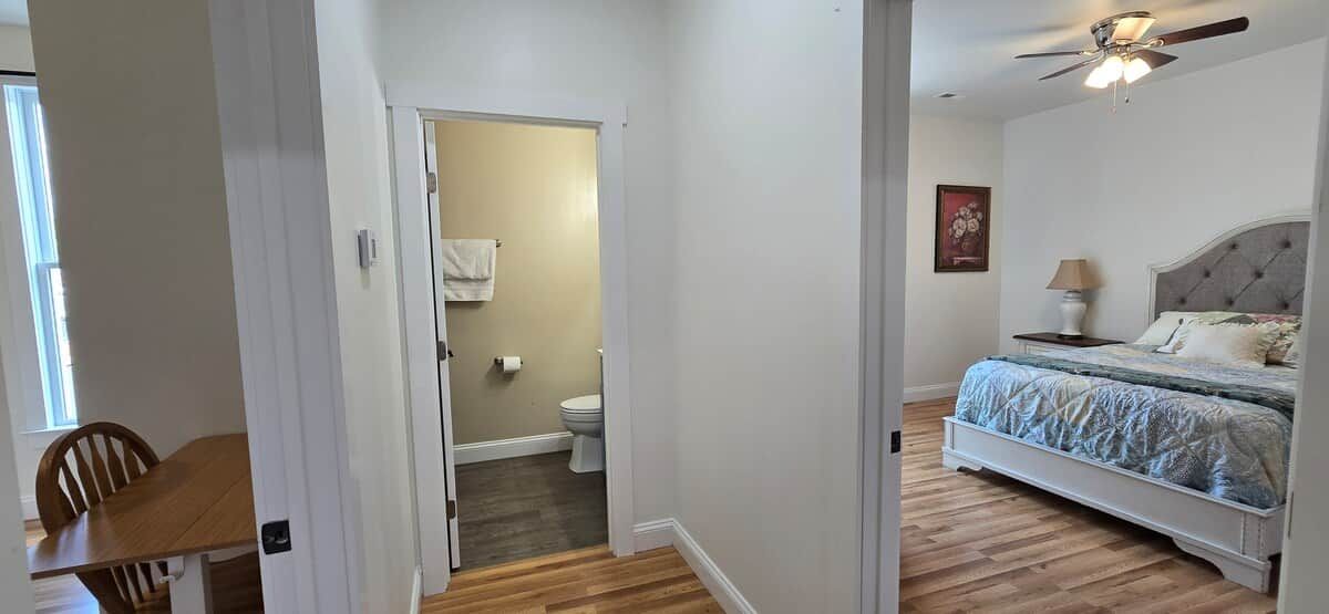 A bedroom with a bed, a nightstand, a ceiling fan, and a bathroom visible through an open door.